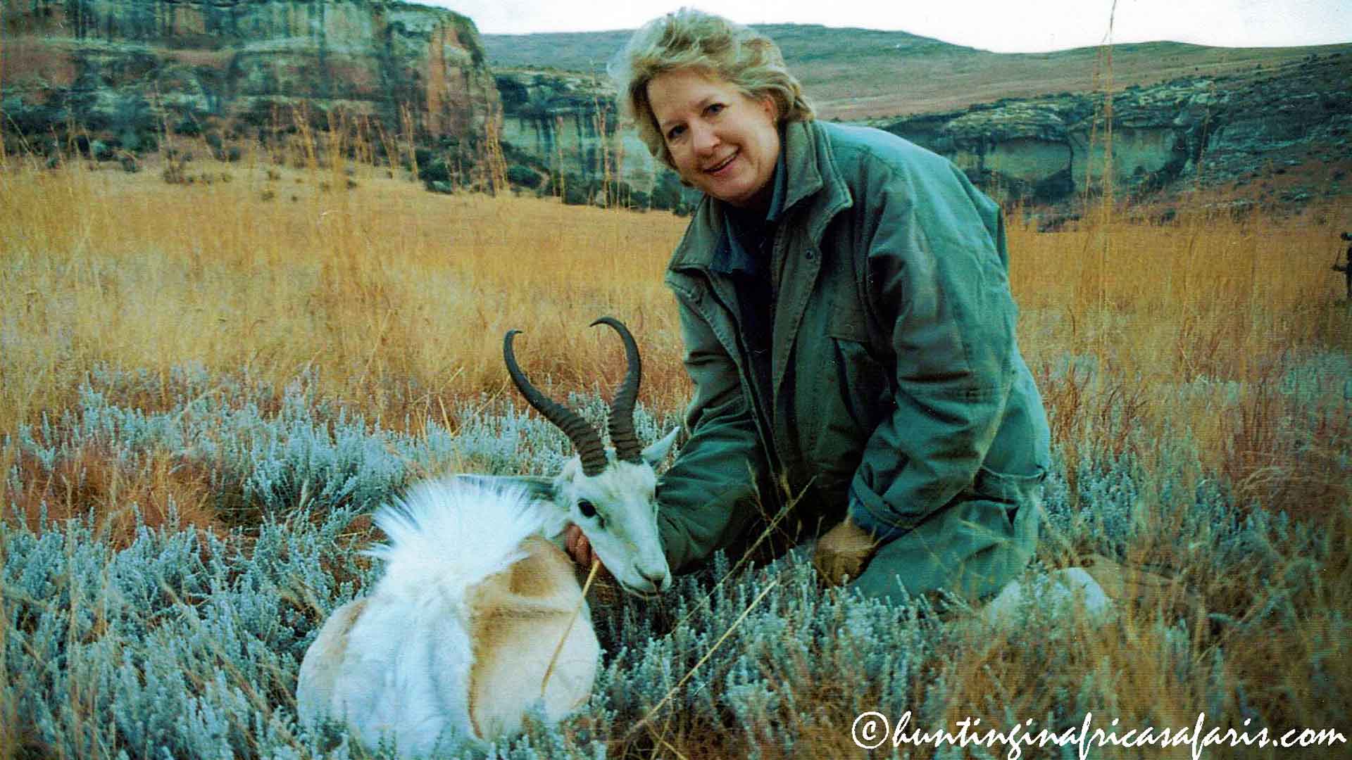 White springbok trophy fees A smiling lady hunter kneeling behind her trophy-size white springbok $text{(Antidorcas marsupialis)}$ in the Eastern Free State, South Africa. The backdrop features a large sandstone mountain and abundant Eucalyptus trees.