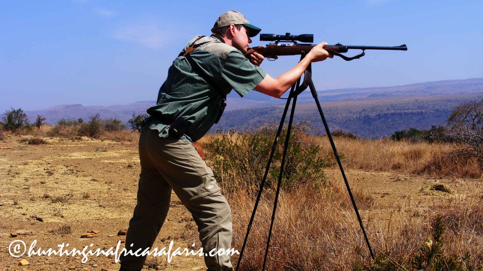 A hunter in a shooting position aiming a .375 H&H rifle over shooting sticks, set against the mountainous landscape of the Ohrigstad region on the Limpopo and Mpumalanga border.